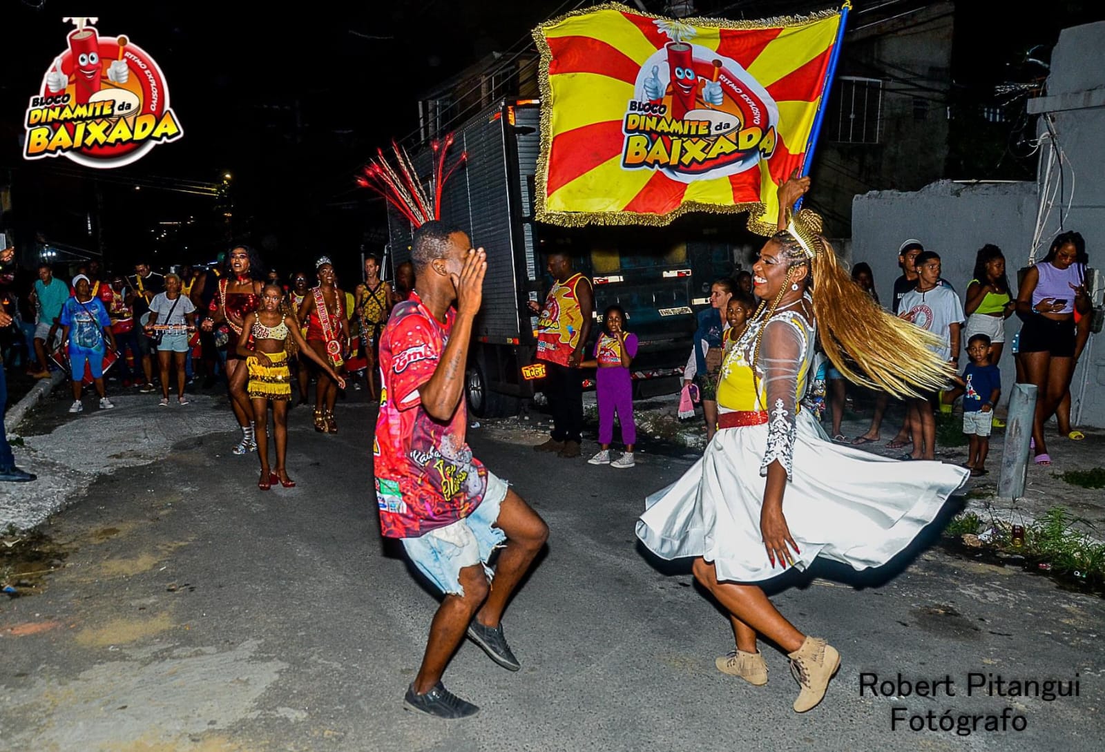 Foliões dançando com o estandarte do Bloco Dinamite da Baixada durante apresentação noturna